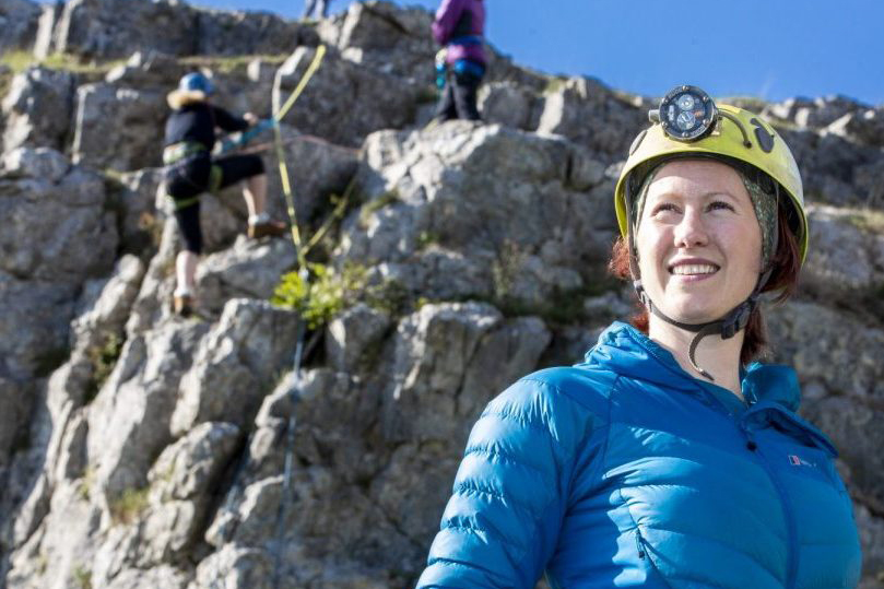 A woman in a blue puffer jacket wearing a helmet with torch stands in front a rocky hill.
